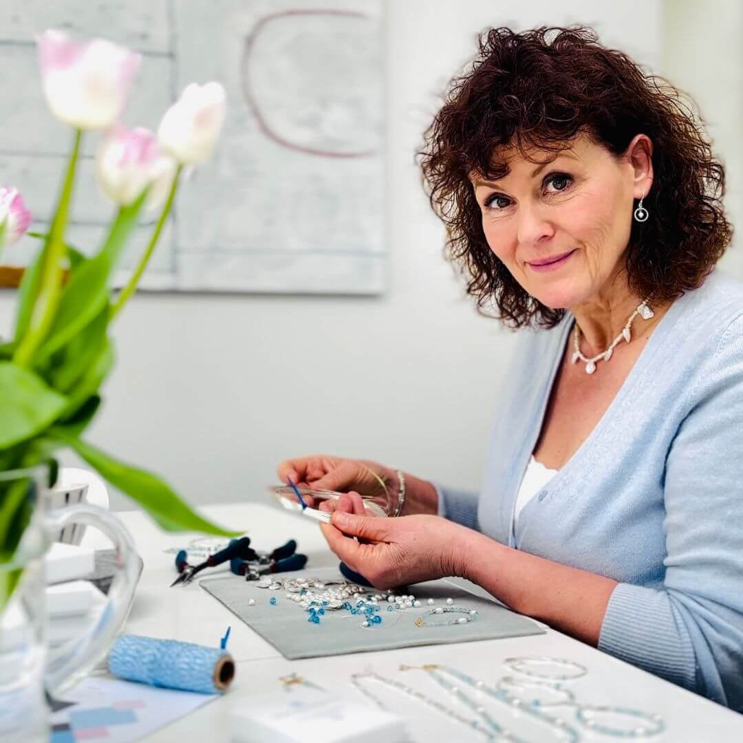 Woman working on jewelry at a table with flowers in the background