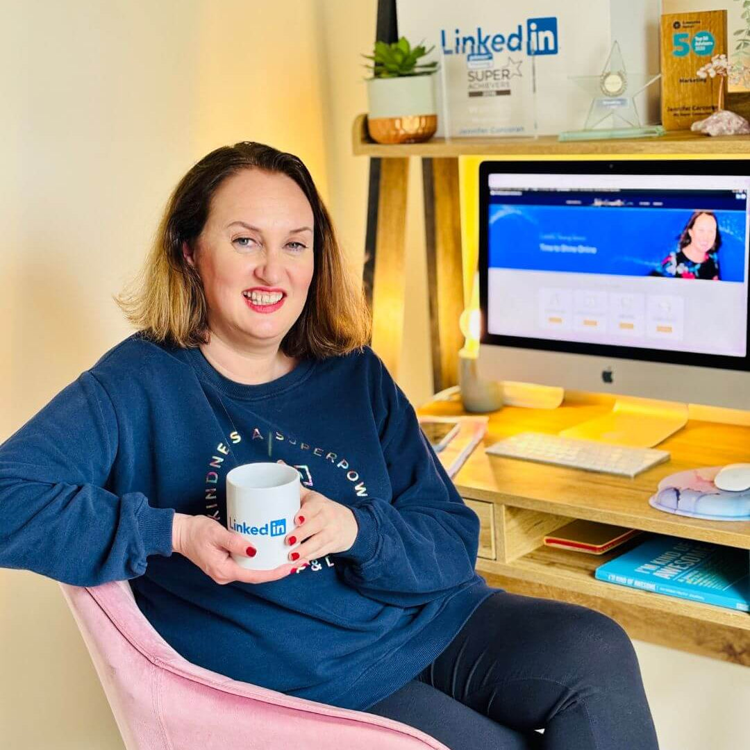 Woman sitting on a pink chair holding a container, with a computer screen displaying LinkedIn in the background.