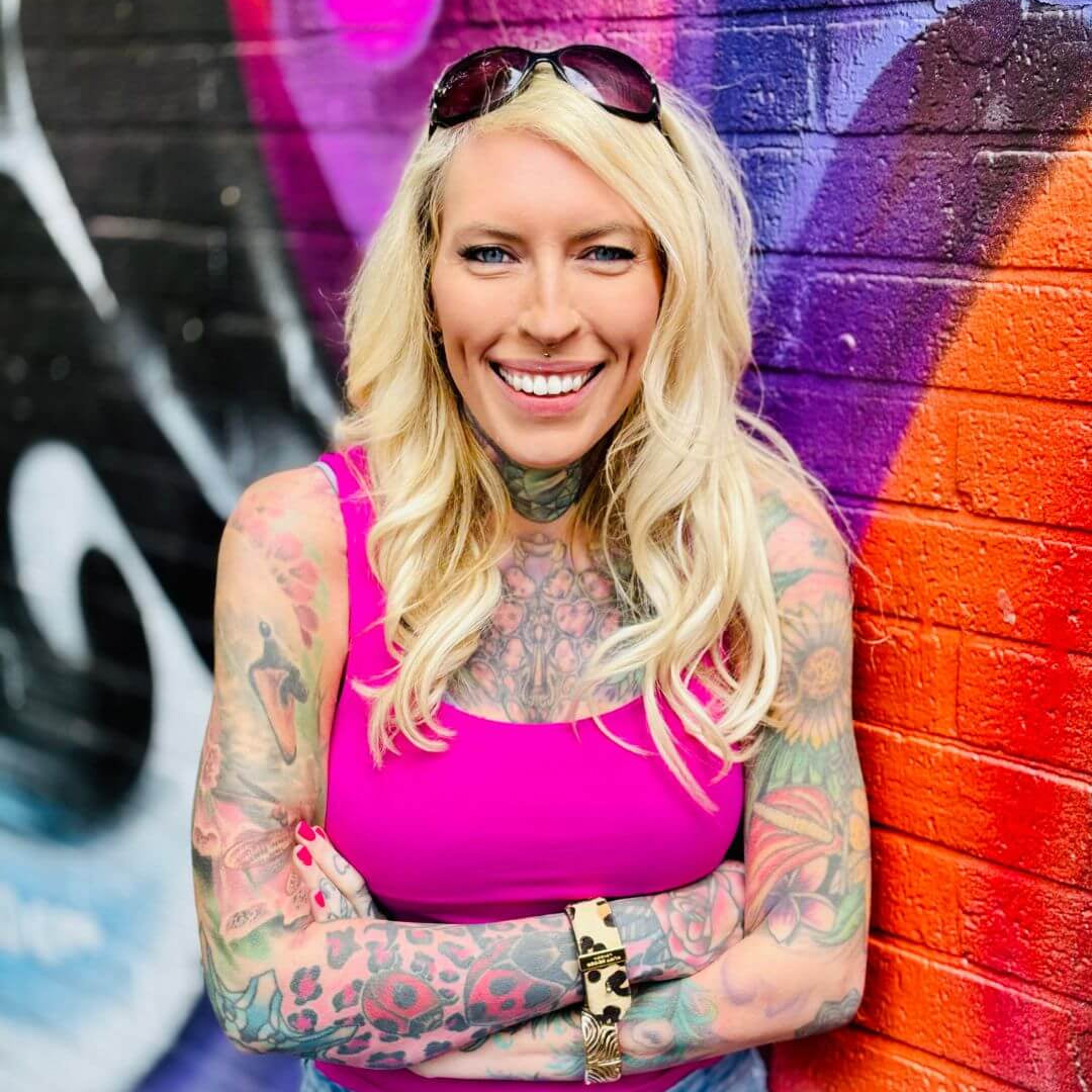 Lea Turner with tattoos wearing a pink top and sunglasses, standing in front of a colourful graffiti wall.