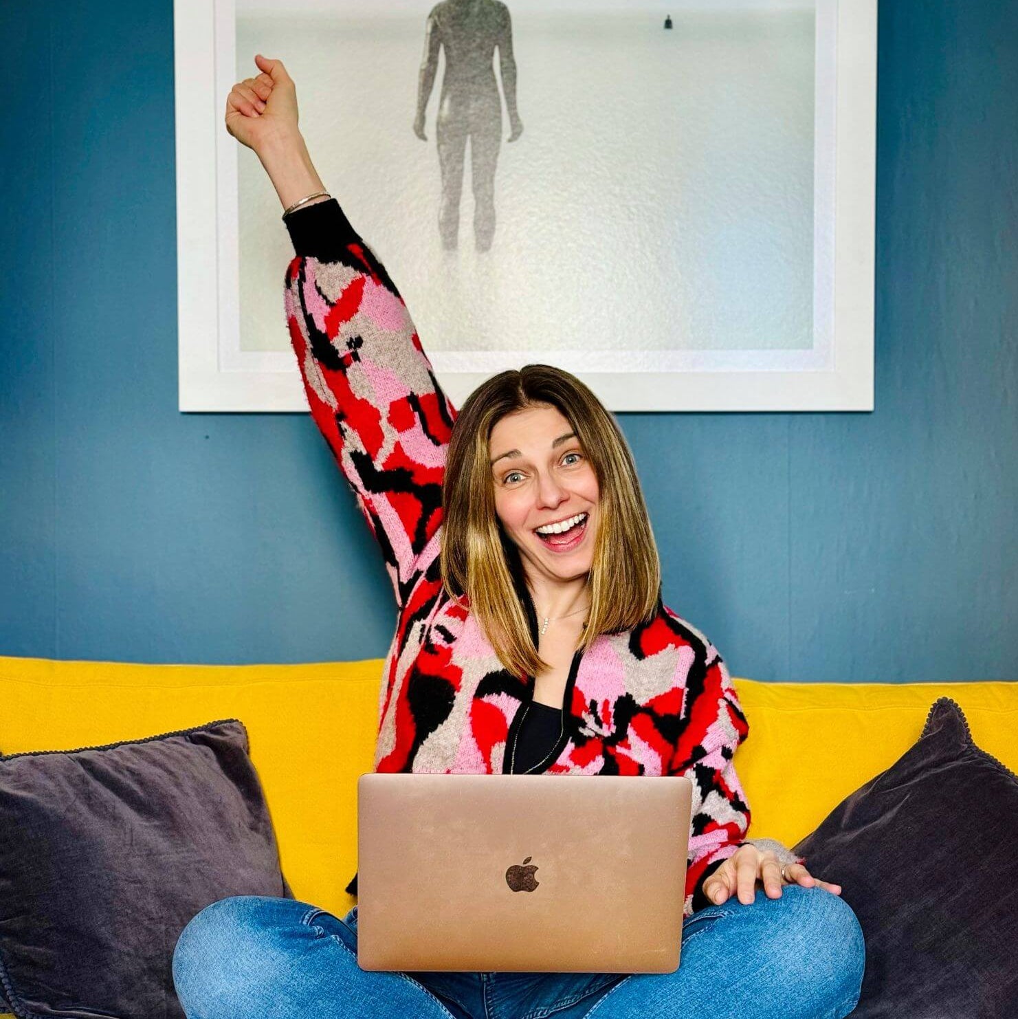Woman sitting on a yellow couch with a laptop, arms raised in excitement, against a blue wall with a framed picture.
