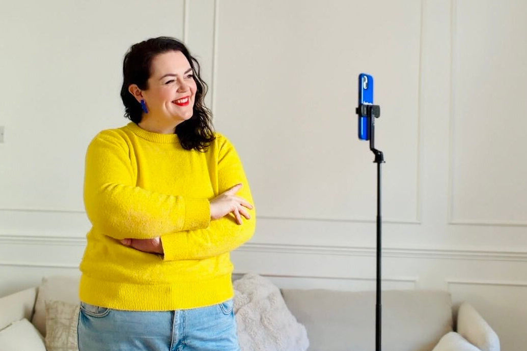 Woman in yellow sweater and jeans standing in a living room with a tripod stand and iphone taking a selfie.